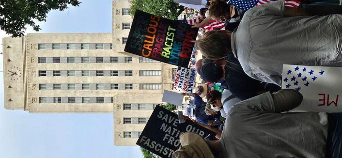 In the foreground, the back of protestors. In the middle, some signs visible reading, "SAVE OUR NATION FROM FASCISM;" "AMERICA THE BEAUTIFUL BORN FROM IMMIGRANTS;" "SUPER CALLOUS FRAGILE RACIST LYING NAZI POTUS." In the background rises Houston City Hall.