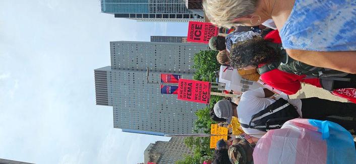 Protestors marching in downtown Houston. Transgender pride flag visible in left foreground. Signs held above protestors' heads read, "I NEED TO BE ABLE TO TELL MY CHILDREN I DID NOT STAY SILENT;" "IF HE CAN GET RID OF FEMA WE CAN ABOLISH ICE;" "ABOLISH ICE RESIST RACISM." A lightpole in the midground has flags hanging that say, "HOU ARE HERE" and "WELCOME TO DOWNTOWN." Buildings rise in the background below a cloudy sky.