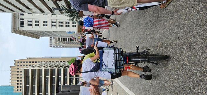 Photo of protestors marching through the streets of downtown Houston. A person walking a bicycle has a sign attached to the back of her bike reading, "Vaccine Save Lives. MAGA KILLS." Another person is draped with an American flag. Multistory buildings rise in the background against a cloudy sky.