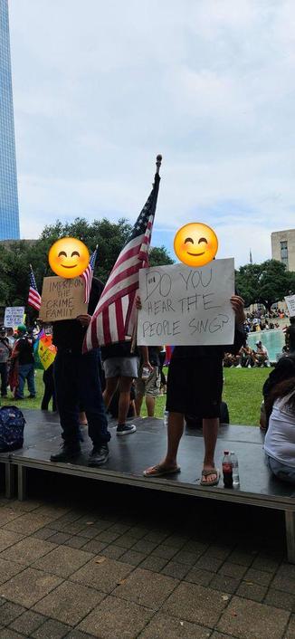 Two people are standing on an elevated platform in a green space in front of Houston City Hall. They are holding signs that read, "IMPRISON THE CRIME FAMILY" AND "DO YOU HEAR THE PEOPLE SING?"