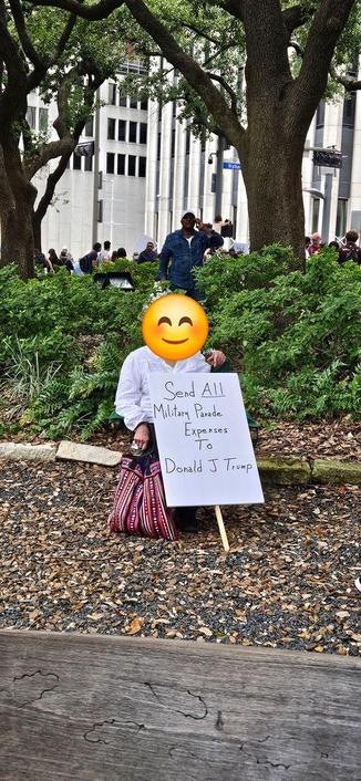 A person seated in a park holds a sign that reads, "Send ALL Military Parade Expenses to Donald J Trump."