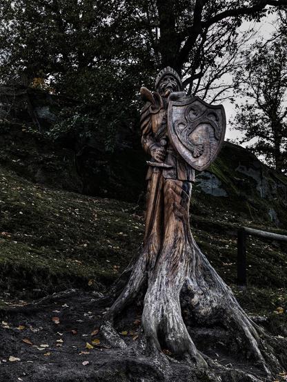 A tall, intricate tree carving depicts a medieval knight as a skeleton, holding a shield and a sword. The knight's armour and helmet are detailed, and the shield features an elaborate design. The carving emerges from the broad, gnarled roots of the tree, which are visible at the base. The setting is a dark, possibly autumnal, woodland with fallen leaves on the ground and trees and rocks in the background.
