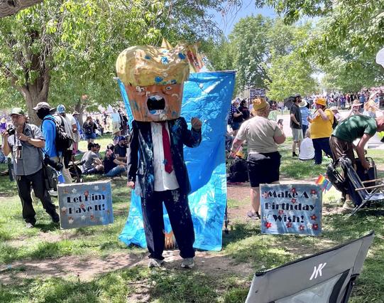 At the Albuquerque No Kings Rally, a man dressed in a costume which includes a blue business suit, a white shirt, and red tie with a gigantic papier mâché head characterizing Donald Trump with an open mouth expression. The man also has lots of cake and icing on his suit. Next to him are signs which read “Let him have it!” And “Make a birthday wish!”