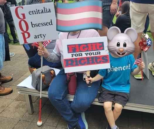 Two people  sitting, holding signs. Their faces are covered with stickers. On the left, me, with my white cane. My sign has red letters, "GOP" down the left side, completed in black to say, "Genitalia Obsessed Party" My head has a Trans flag covering my face. The grandkid has a blue shirt with "Science" in white. Their sign says, "Fight for your Rights" in all caps. Their face has a cute mouse sticker covering it.