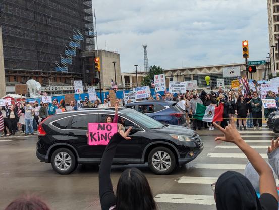 Some of the cars driving by and holding signs. This particular one is pink and reeds no Kings. The passenger in the car that is holding the sign is flashing a peace sign with her hand. 

Other protesters line the roads and cheer her on as she cheers us on.