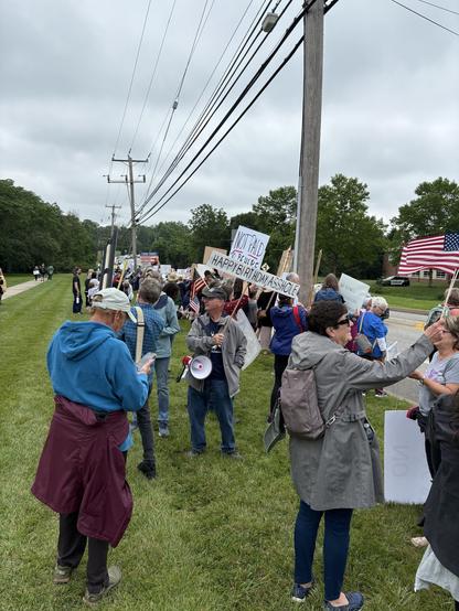 A group of protesters stands along a grassy area next to a road, holding various signs. Some signs display messages such as "NOT PAID" and "HAPPY BIRTHDAY ASSHOLE." The crowd includes a diverse mix of individuals.