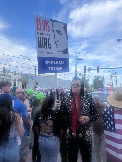 A crowd gathers at a protest with a person dressed as Elvis holding a sign that reads "ELVIS is the only KING in Las Vegas" and "IMPEACH TRUMP." The background features diverse attendees.