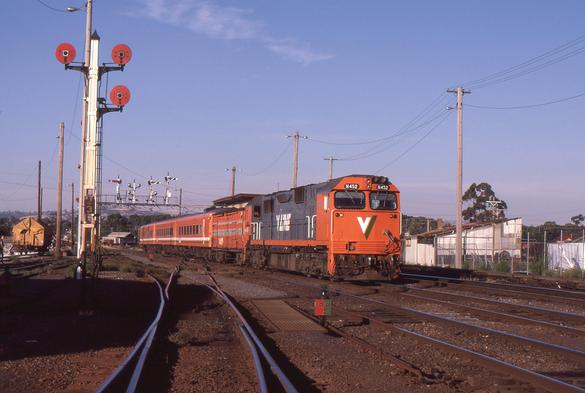 An orange and grey VLine liveried N class narrow carbody/cab style diesel locomotive hauling a tangerine liveried passenger train comprised of a baggage van plus three passenger carriages arrives into a station yard.  A tall post with three disc signals stands at left, a ground level points indicator is at lower right, and a gantry of lower quadrant semaphore signals spans the tracks in the background.  A yellow liveried, tarpaulin-covered GY wagon is sitting in the yard at left.