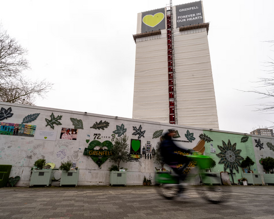 Looking at the Grenfell tower, surrounded by a wall with slogans and a bicycle passing by in front of