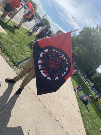 A demonstrator holds a red, black, and white flag with the IWW logo in its center. 

The logo is surrounded by black silhouettes of cats, whose white eyes can be prominently seen. 

The IWW was well known for its "wildcat strikes" that occurred spontaneously, without prior permission from union leadership.
