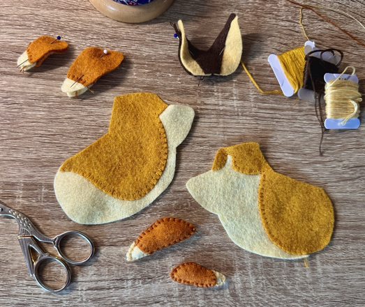 Picture of a work in progress set on my desk. Half assembled pieces of orange, cream and brown-coloured felt slowly taking the shape of a future plushie. A few assorted threads, some pins, and embroidery scissors are placed around it.