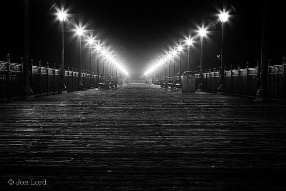This Is A Very Dark And Grungy Black And White Street Photo, Without People, In Landscape Format. Stretching Upwards From The Base To A Little Over Half Way Up In The Frame Is A Surface Made Up Of Rough And Weathered Planks Of Wood Laid At Right Angles To The Camera. From Either Side Of The Image And Converging Inwards Towards The Middle Are Two Parallel Rows (Though They Appear To Converge) Of Metal Railings. Inset From Those Railings Are A Line Of Metal Street Lights, Fifteen To Twenty, That Again Appear To Converge Towards The Centre. The Tops Of Those Lights Have An Eight Point Star Glow, In Addition To Ever So Slightly Illuminating The Warm And Humid Air. To Add Some Municipal Ambiance To The Scene There Are Some Outward Facing Benches And A Few Wast Bins. 
The Location: Pier 7, San Francisco (2012)
