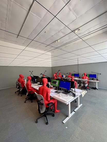 Room staged as an architecture office. Red mannequins sit at white desks with computer screens, lamps, and scattered papers. The mannequins represent architects working outside traditional building practice. Monitors show red, green, and blue-themed content symbolizing participants, projects, and student surveys. The space evokes a once-prestigious but now abandoned studio.