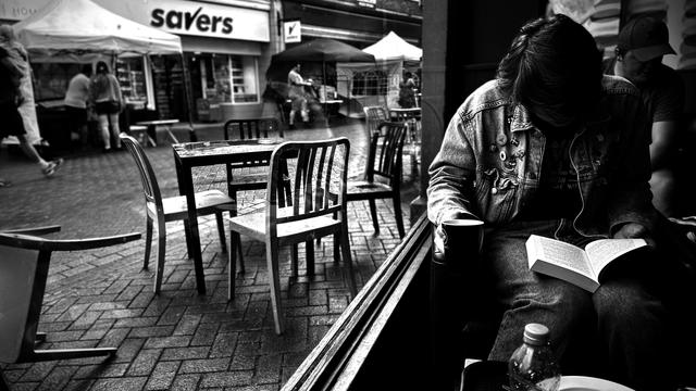 A youth in denim sits in a coffee shop, mug in one hand and a book in the other. Their head is down and they are intend on reading. To the left is a street scene of empty chairs and tables