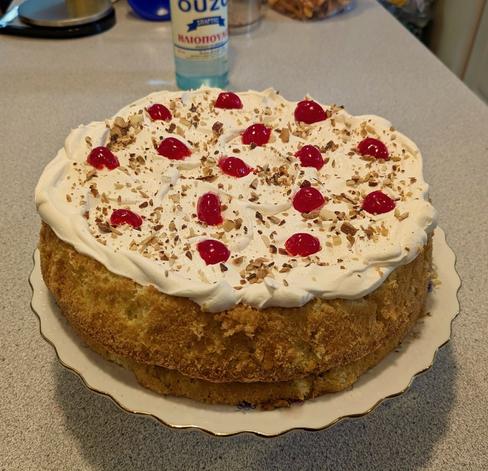 A double layer sponge cake infused with a sweetened Ouzo (Anise flavored Greek liquor and whole lactose free milk) topped with Cool Whip, toasted almonds chopped in small bits, and fifteen halved maraschino bright artificially red cherries. Unseen is a chocolate buttercream filling. Blurred in the background is a bottle of Greek Ouzo from Sparti, Greece. A kitchen scale and other blurred items from the kitchen island (grey formica).
