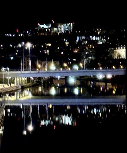Nächtlicher Blick auf die Saar mit einer Brücke und vielen Straßenlaternen, die leuchten, dahinter geht es den Berg hoch, auf dessen Kappe zwei erleuchtete Hochhäuser stehen.