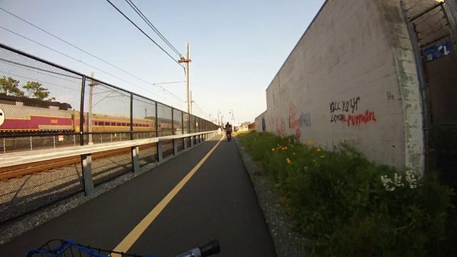 a picture from a handlebar camera on a bicycle. the bike is on an asphalt path between train tracks and commercial buildings. on the left, a train. on the right, a building with graffiti reading "KILL YOUR LOCAL PUPPYGIRL"