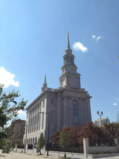 The image features a tall, multi-story building with a distinct spire and a golden statue at the top. The architecture appears to be classical with a light-colored exterior. Surrounding the building are trees and plants, and the sky is clear with some clouds. This structure resembles a temple or a significant religious building, often characterized by ornate details and a commanding presence.

Image Credits: Wikimedia / JonRidinger / CC BY-SA 4.0