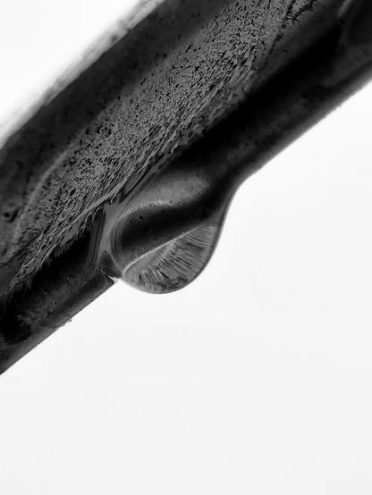 Detailaufnahme in Schwarzweiß von einem Regentropfen, der an einer Metalllamelle einer Außenjalousie hängt.
