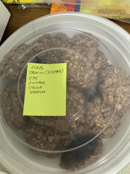 Oatmeal cocoa blobs in a plastic container with a Post-it on it with writing saying oats, tahini, egg, sugar, cocoa, vanilla