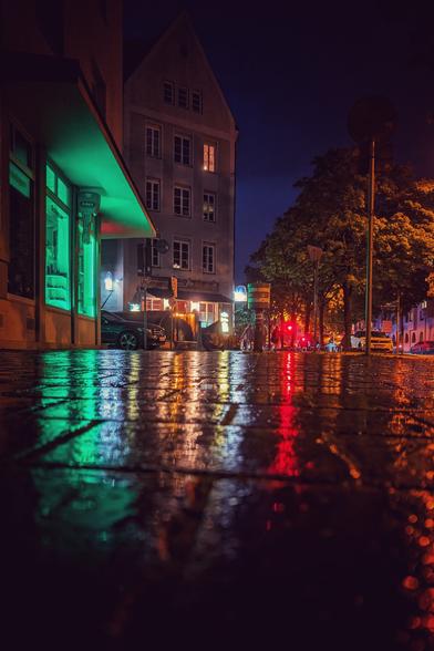 Das Bild zeigt eine nächtliche Straßenszene mit regennassem Pflaster, das die farbenfrohen Lichter der Umgebung reflektiert. Die Perspektive ist niedrig, fast bodennah, sodass die glänzende Oberfläche des Straßenbelags besonders hervorsticht. Links gibt es ein Gebäude mit einem auffälligen grünen Licht, das aus einem Fenster oder einer Tür strahlt. Im Hintergrund erstrecken sich weitere Gebäude und Bäume, sanft erleuchtet von Straßenlaternen und anderen Lichtquellen. Die Farben im Bild sind überwiegend rot, gelb und grün.
