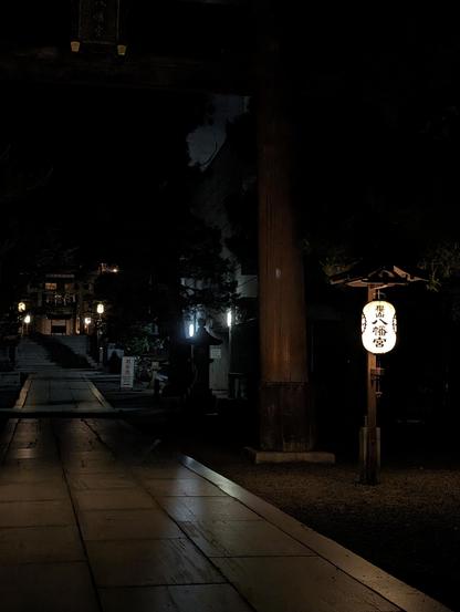A Japanese lantern glowing in the middle of the night within the grounds of a shrine. The place is empty and quiet. The torii right column can be seen in the forefront, with the shrine's main hall in the background.