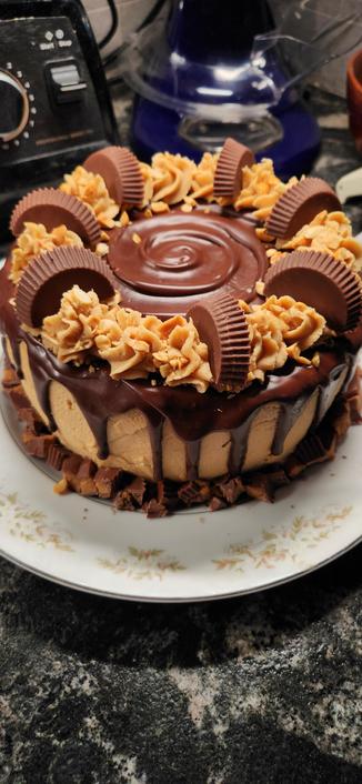 Gluten free chocolate layer cake with peanut butter frosting. There's a swirl of chocolate ganache in the center & drips of it along the sides. Peanut butter cups cut in half are  on the frosting on the top.