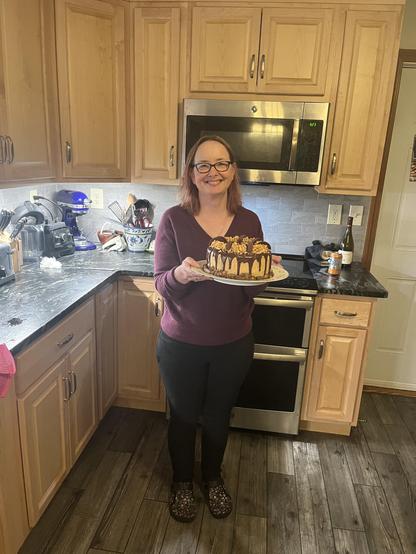 A white middle aged woman standing in a kitchen holding a plate with a cake on it.