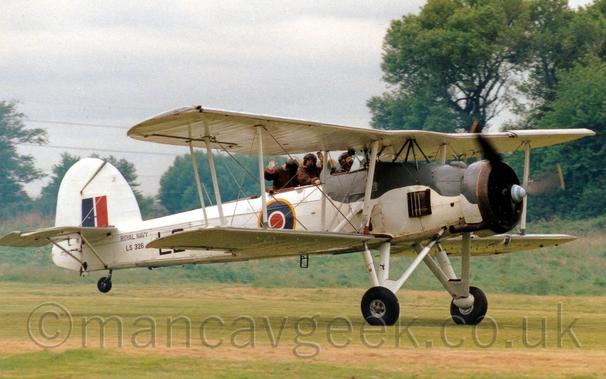 Side view of a large, single-engined bi-plane rolling along a grass runway with it's tail-wheel raised into a flying position.
The plane is mostly white, with the upper forward fuselage and engine cowling painted black, and a blue, white, and red roundel outlined in yellow under the rear cockpit in the middle fuselage.
There are small "Royal Navy" titles on the rear fuselage, above the serial "LS326".
There is a red and blue fin flash on the tail.
There are 3 people visible in the 2 open cockpits on top of the plane, all bundled up in thick leather, fur-lined flying jackets and leather flying jackets - the pilot in the front cockpit, facing forward and concentrating on flying, as he should do, and 2 crew in the rear who are waving at the crowd.
Short green grass fills the foreground, with trees lining the airfield perimeter in the background, under cloudy grey skies.