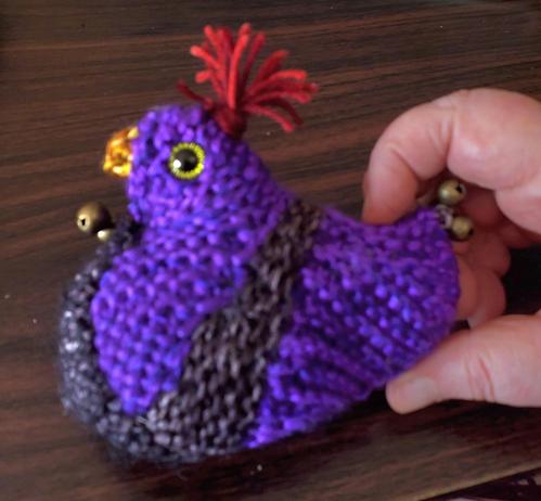 Side view of chickie, purple with black stripe and underbelly, red comb, yellow beak, green eyes, 3 bells at neck and tail each