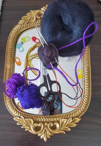 My latest knitting set-up, viewed from above - a small, gilt, mirror-backed tray, the gilt sides forming a shallow lip, on top are miscellaneous scrap yarns, scissors, stitch holders, a circular needle