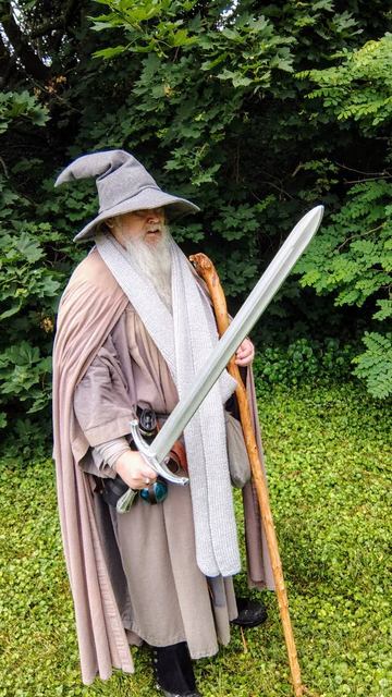 Middle aged man with long grey beard and Gandalf style outfit with hat, scarf, and staff, wielding a Glamdring replica in front of some green underbrush.