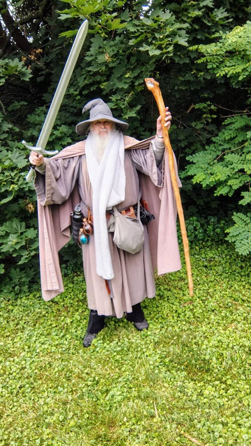 Middle aged man with long grey beard and Gandalf style outfit with hat, scarf, and staff, wielding a Glamdring replica in front of some green underbrush.