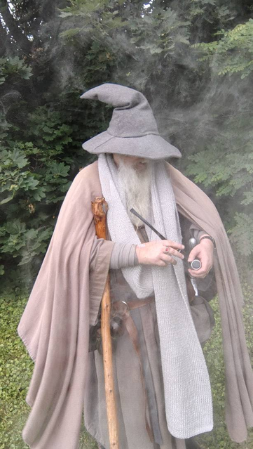 Middle aged man with long grey beard and Gandalf style outfit with hat, scarf, and staff, holding a black vape pipe with smoke in the foreground