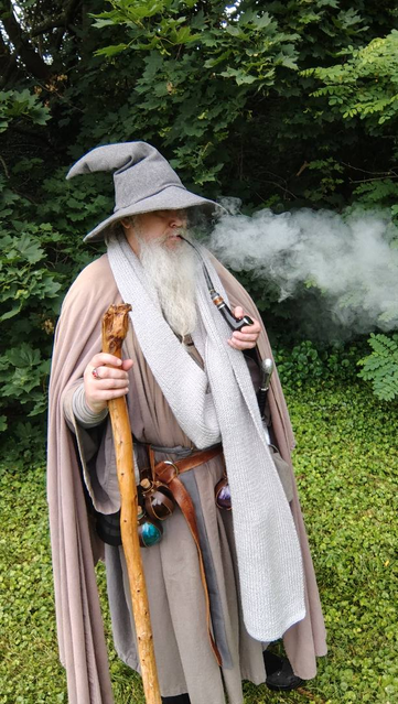 Middle aged man with long grey beard and Gandalf style outfit with hat, scarf, and staff, smoking a black vape pipe.