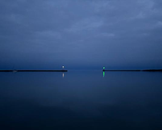 Looking out at the harbor after dusk. Lights at the end of the jetty are blinking on. Dark blue of a calm Lake Superior gradually blends into the cloudy gray of an overcast sky.