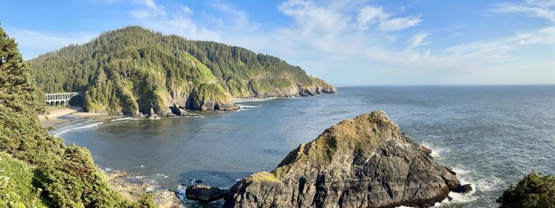 A panoramic view of a coastal landscape featuring a rocky shoreline, gentle waves, and lush green hills. A bridge arches over a river leading to a beach, with trees lining the cliffs and clear skies overhead.