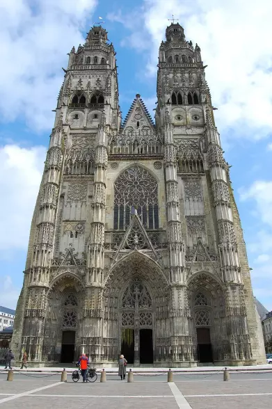 The image depicts a grand Gothic cathedral with impressive towers and intricate stonework. The façade features detailed sculptures and stained glass windows, typical of Gothic architecture. There are people in the foreground, including someone on a bicycle and another person walking, which adds scale to the scene. The sky appears to be partly cloudy.

Image Credits: Wikimedia / Goldmund100 / CC BY-SA 3.0