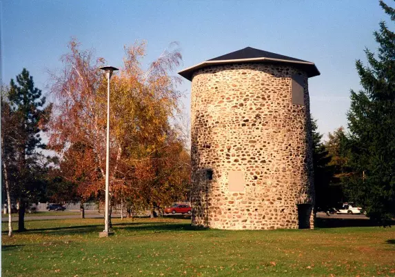 The image shows a stone structure that appears to be a cylindrical tower, likely made of a mix of large and small stones. It has a dark, possibly conical roof and is situated in an open area with grass. Surrounding the tower are trees, some of which show autumn foliage, indicating seasonal change. There is also a streetlight nearby and a car visible in the background. The overall scene suggests a park-like setting.

Image Credits: Wikimedia / René Beaudoin / CC BY-SA 3.0