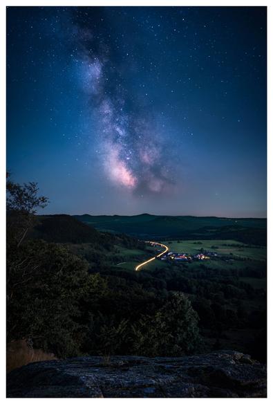 The photo was taken from a rock and shows a valley at night. Above the valley is a bluish glowing Milky Way. A small village can be seen in the valley, which is brightly lit. On the road to the village there is a trail of light from a moving car.