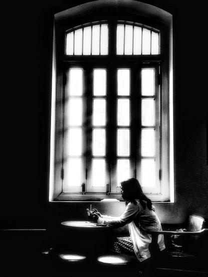 Black and white photo of a giant window illuminating a silhouette of a person using their phone at a table in a coffee shop