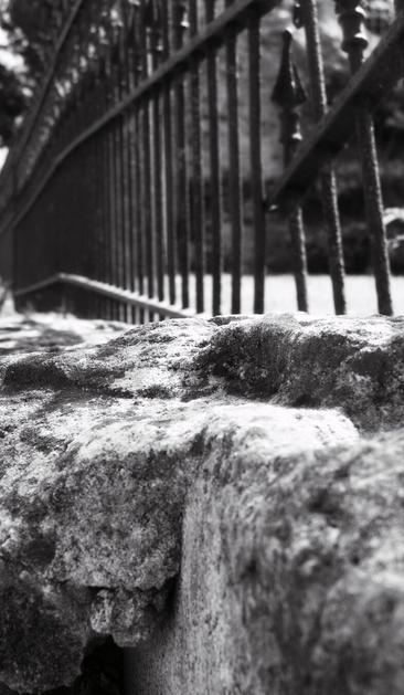 A black and white photo showing a low angle view of a stone wall with a black metal fence.