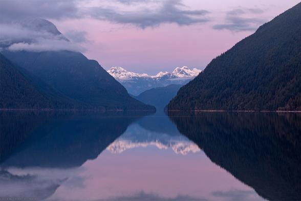 A reflection of mountains in a lake after sunset - with a purple sky.  Mountains on the left and right are close by, covered in trees, and relatively low elevation.  In the middle of the frame are more distant, rocky, mountains with some fresh snow on them.