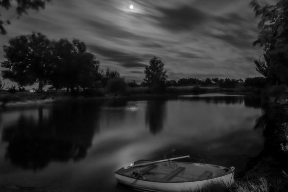 Black and white night photograph showing a raft with a boat in the foreground and a night sky with clouds and a full moon between them.