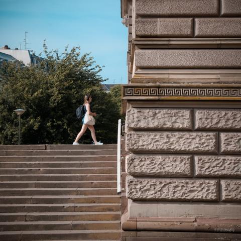 A woman with a backpack walks rightward, preparing to enter a large old building made of textured stonework and classical architectural details. She is walking on the forecourt of the building, which is located at the top of some steps visible in the lower left corner. The building occupies the right side of the photo.

Urban-style vibrant colours edit