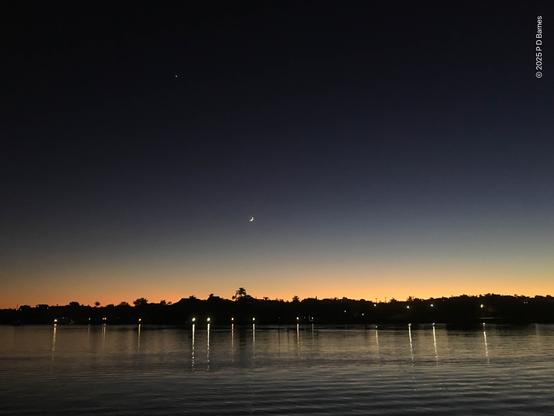 Looking across a calm Brisbane river at dawn. On the opposite bank the land is still dark, lights on the riverwalk reflecting in the river, trees silhouetted against the early orange glow just creeping over the horizon. Higher up a small crescent moon hangs in a slate-blue, almost black-ink sky.