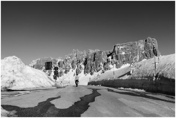 A black and white photo taken in the Dolomites in northern Italy. The subject of the photo is a craggy rock face and in the foreground there is a asphalt road flanked with giant snow banks leading toward the mountain.
