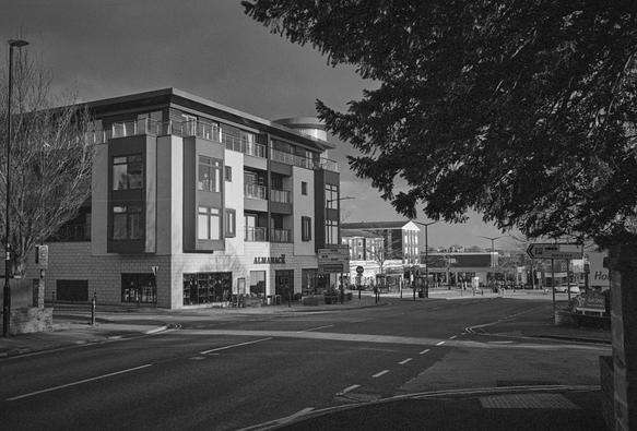 Black and white photo of a modern apartment building with a bar on the ground floor, seen under the branches of a yew tree. The sky above the building is dark and threatening.