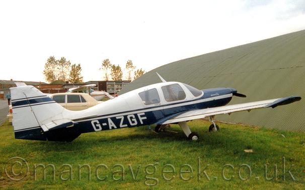 Side view of a single engined light aircraft parked on grass facing to the right.
The plane is mostly dark blue, with white upper surfaces, a white stripe running along the body, and the white registration "G-AZGF" on the lower rear fuselage.
There is small white text reading "Beagle Pup" on the engine cowling, and small black text reading "Pup 150" under the cockpit windows.
A blue stripe also runs along the top of the tail.
Green grass fills most of the foreground, with the sharply-angled green metal side wall of a hangar dominating the background on the right, with a sandy brown car, a white light aircraft, and a couple of brown buildings in the background on the left, all under bright but hazy sky.