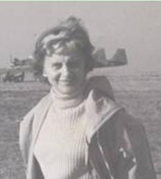 Irena Kempówna in polo neck and anorak with hood down standing smiling on airfield with glider in background